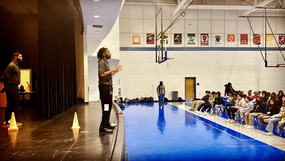 A Power of Water Foundation staff member leads a coaching session in a school gymnasium filled with students, highlighting personalized support for families and emotional well-being education.